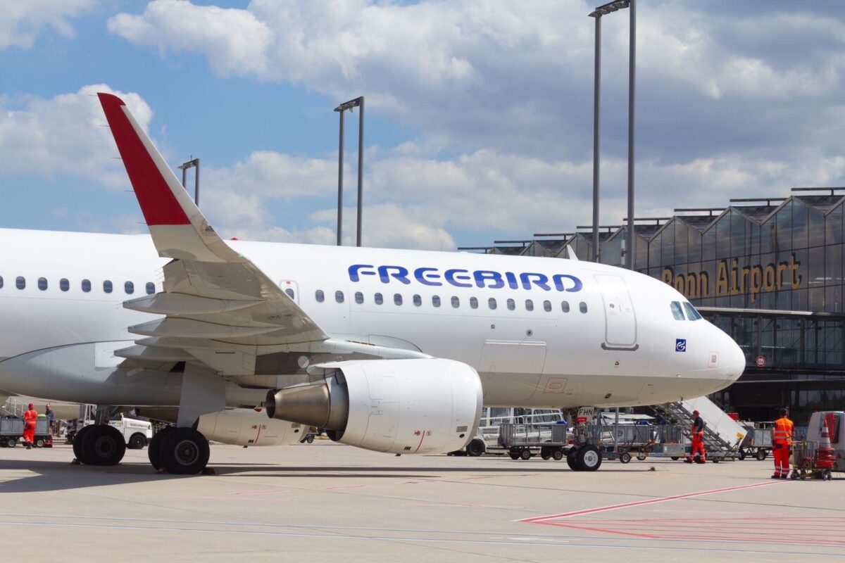 Eine Maschine von Freebird Airlines steht auf dem Vorfeld vom Flughafen Köln/Bonn bei blauem Himmel. Zu sehen ist die eine Seite der Tragflächen, eine Turbine und die blaue Aufschrift auf dem Flugzeug "FREEBIRD". Im Hintergrund ist am Flughafengebäude ide Aufschrift "Bonn Airport" zu sehen.