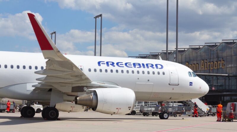 Eine Maschine von Freebird Airlines steht auf dem Vorfeld vom Flughafen Köln/Bonn bei blauem Himmel. Zu sehen ist die eine Seite der Tragflächen, eine Turbine und die blaue Aufschrift auf dem Flugzeug "FREEBIRD". Im Hintergrund ist am Flughafengebäude ide Aufschrift "Bonn Airport" zu sehen.