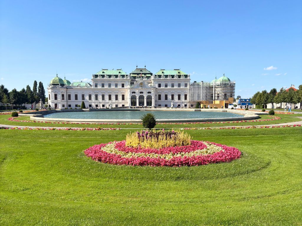 Im Hintergrund ist das Schloss Belvedere zu sehen. Im Vordergrund ein angelegter See und eine Blumenwiese.