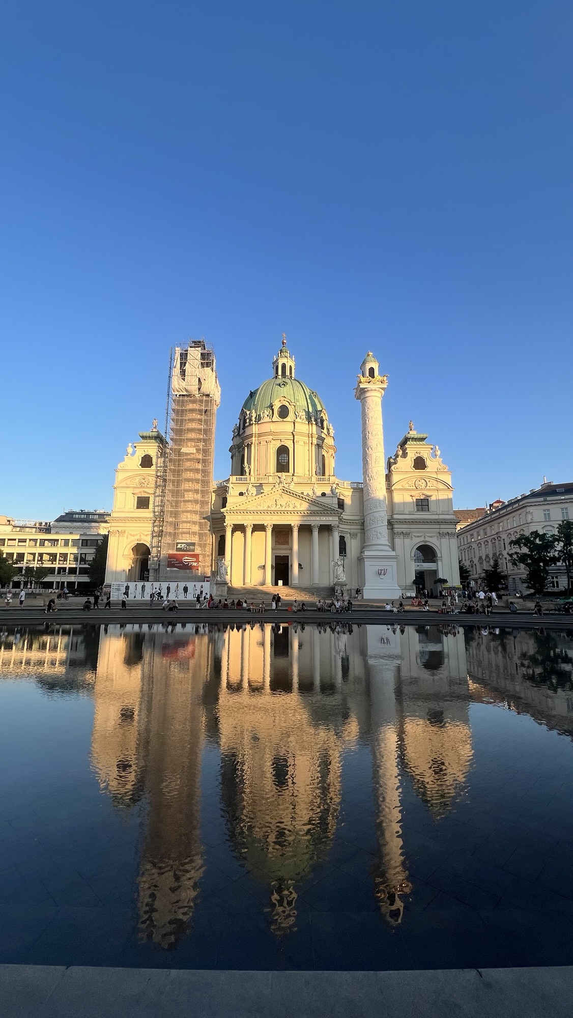 Die Karlskirche spiegelt sich in der Abendsonne im Wasser. Über ihr erstrahlt der himmelblaue Himmel ohne Wolken.